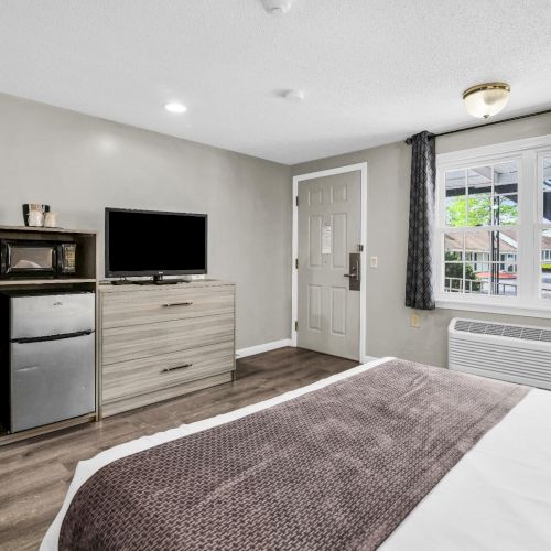 A tidy bedroom with a flat-screen TV on a dresser, a mini fridge, a window with curtains, and a wall AC unit.