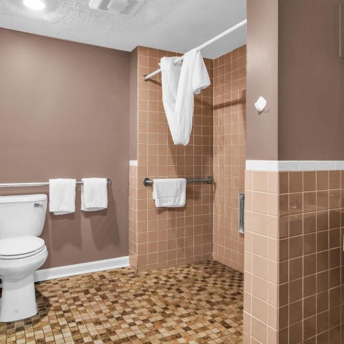 A hotel bathroom with toilet, sink, tiled shower, hanging towels, and safety grab bars.