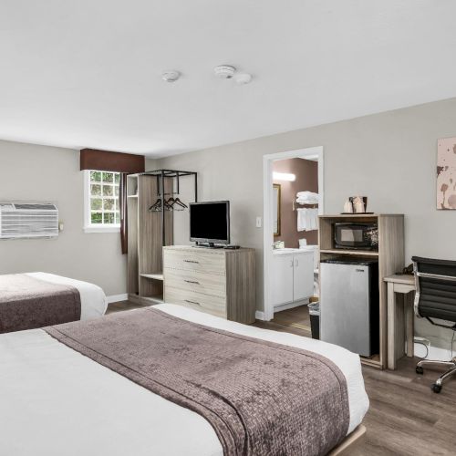 A clean hotel room with two beds, a TV, desk, chair, microwave, mini-fridge area, window AC, and wall art hanging on the walls.