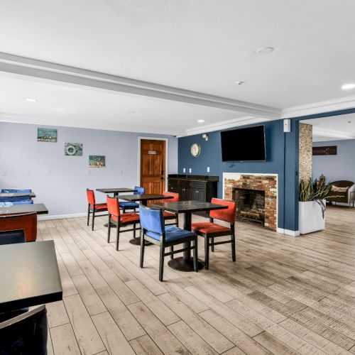 Bright dining lounge with tables and colorful chairs, a brick fireplace, flat-screen TV, and large windows letting in daylight.