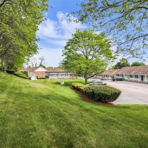 A sunny campus scene with manicured lawns, leafy trees, and low buildings beside a parking lot and walkway.