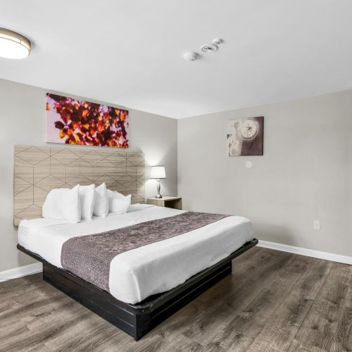 A modern bedroom with a made bed, lamps, framed art, ceiling lights, and a desk area in view.