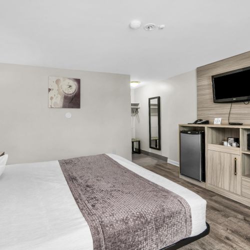 A hotel room with a neatly made bed, bedside lamp, wall art, and a TV and kitchenette cabinet on the right.