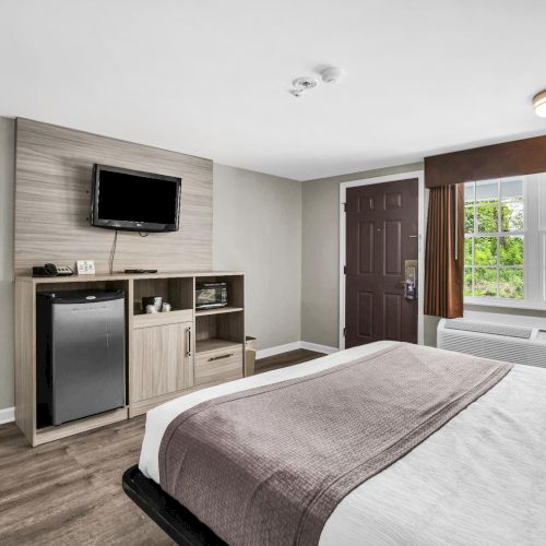 A neat hotel room with a bed, flat-screen TV and mini fridge cabinet, plus a window with curtains and an overhead light.