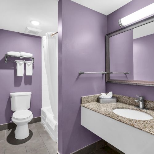 Bright bathroom with purple walls: toilet, granite vanity sink, wall mirror, and neatly folded towels.