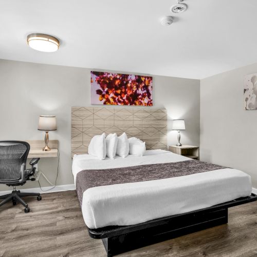 Modern bedroom with a made bed, lamps, wall art, and an office chair by a window.