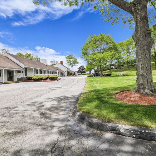 A sunny driveway lined with white building units, manicured lawns, and tall trees under a blue sky with clouds.