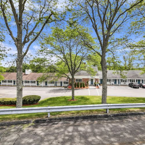 A sunny parking lot framed by leafy trees in front of a long brick-and-white building with a few parked cars.