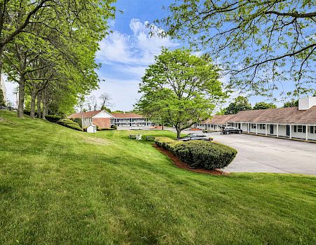 A sunny, well-kept college campus or motel exterior with green lawns, trees, and low-rise buildings lining a driveway. Top it.