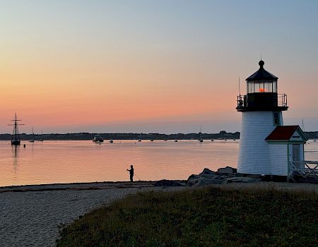 A serene sunset over a calm bay with a small white lighthouse, a lone fisherman on the shore, and gentle colors reflecting on the water.